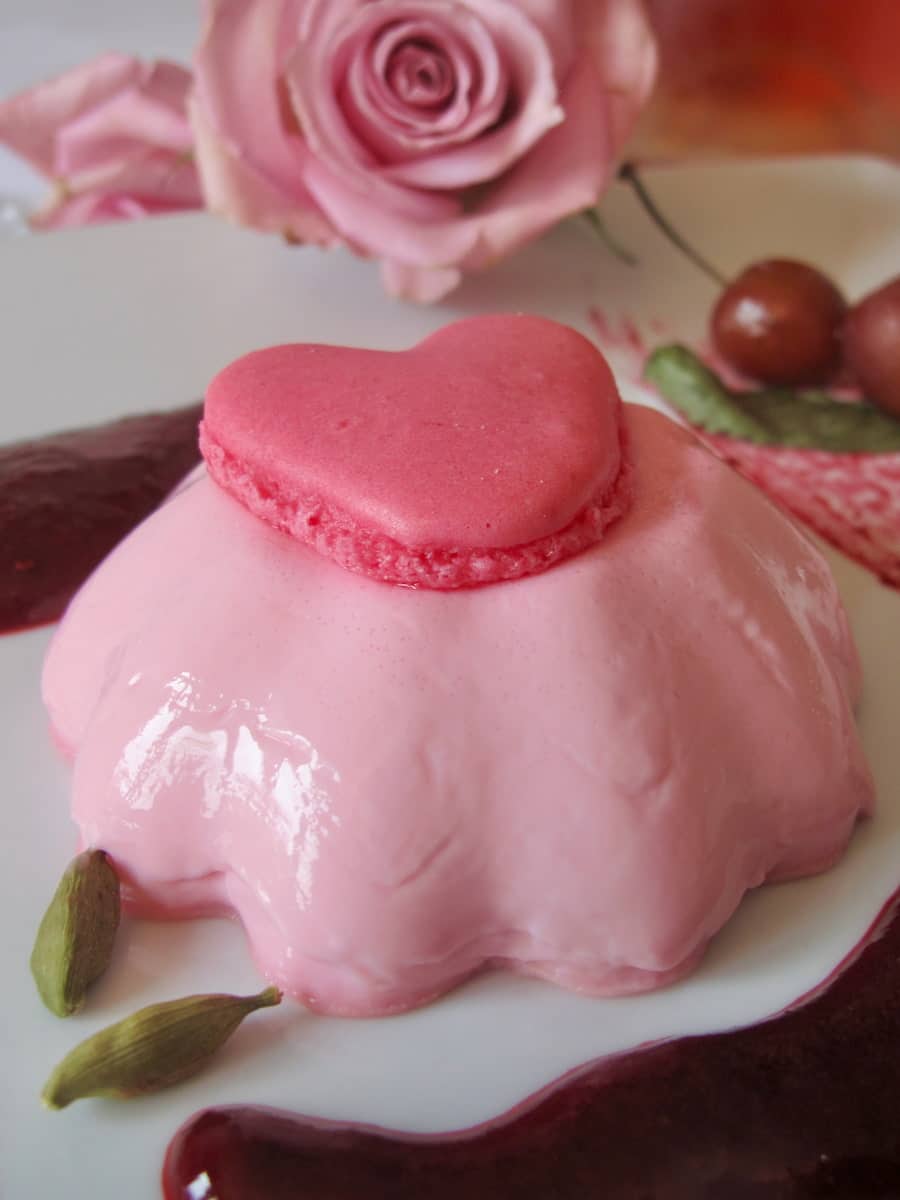 pink fluted dessert topped with a love heart macaron shell surrounded by dark cherry sauce and 2 cardamom pods next to a rose