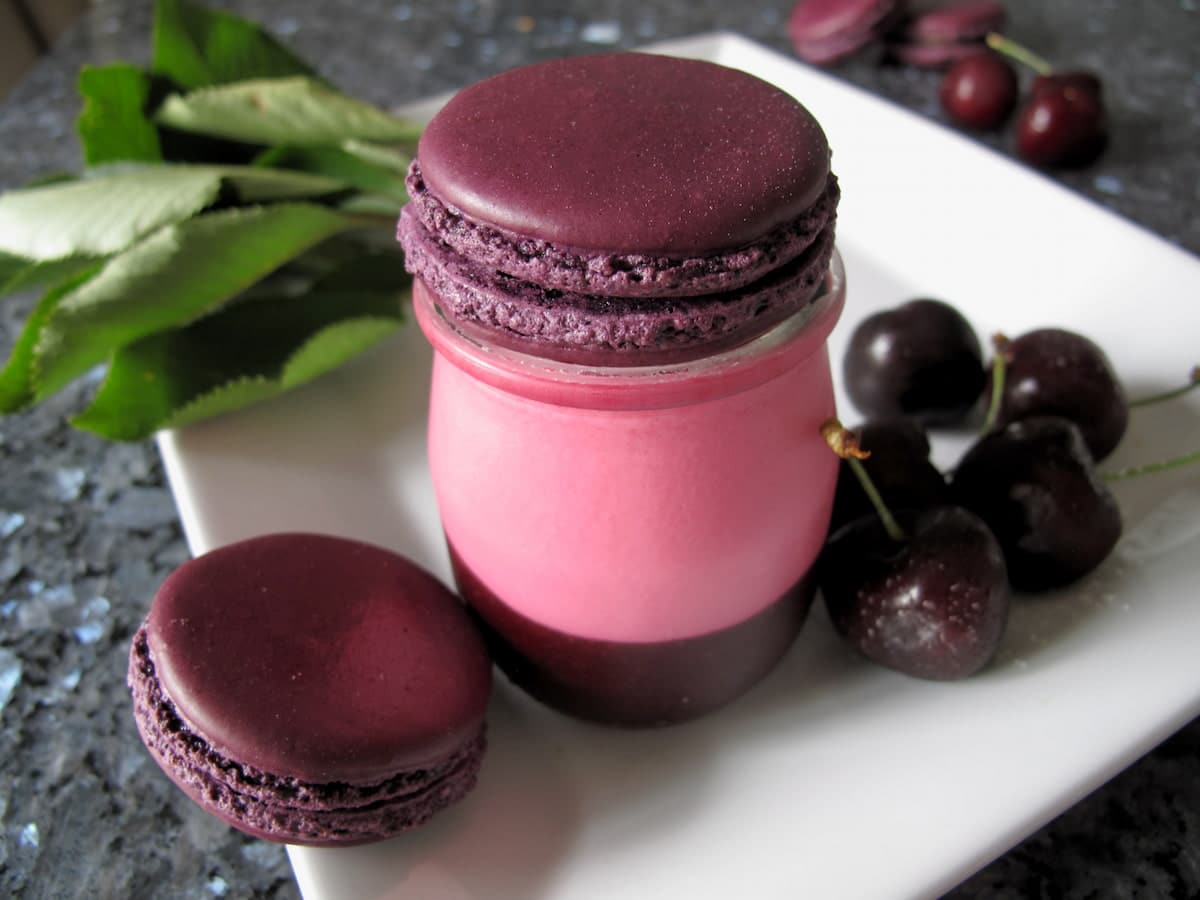 pink and dark cherry coloured filled yoghurt pot next to dark cherries and macarons