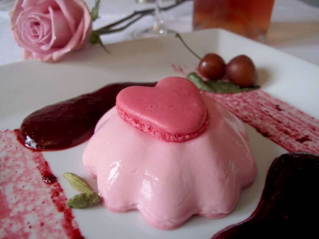 fluted creamy pink dessert topped with a love heart macaron shell surrounded by cherries, cardamom and a rose