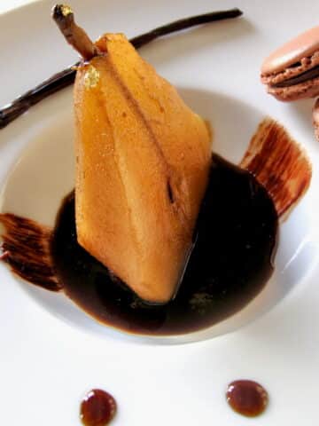 elegant dessert plate with half a poached pear sitting in a puddle of vanilla and coffee syrup with chocolate macarons