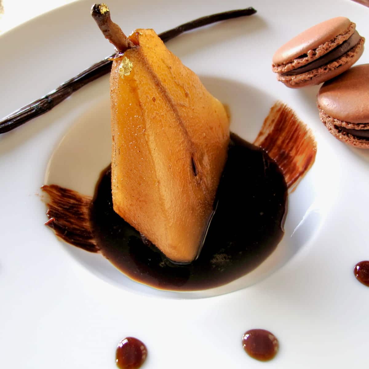 elegant dessert plate with half a poached pear sitting in a puddle of vanilla and coffee syrup with chocolate macarons