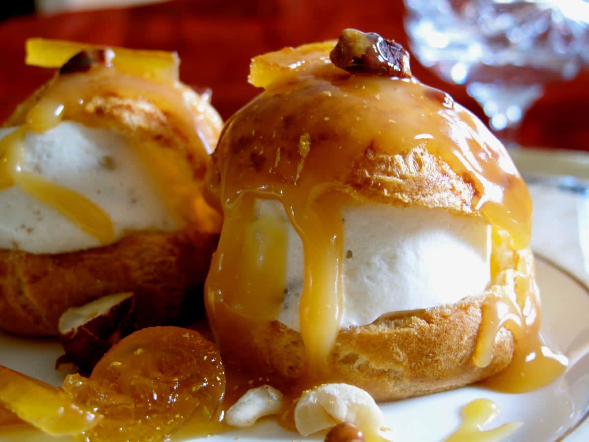 2 profiteroles filled with ice cream and topped with runny golden sauce surrounded by candied fruits and toasted nuts