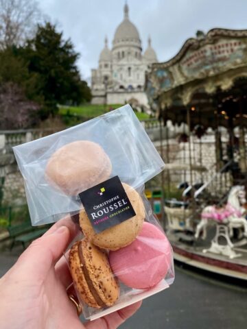 holding a pack of Parisian macarons in front of a carousel and Sacré Coeur basilica in Paris