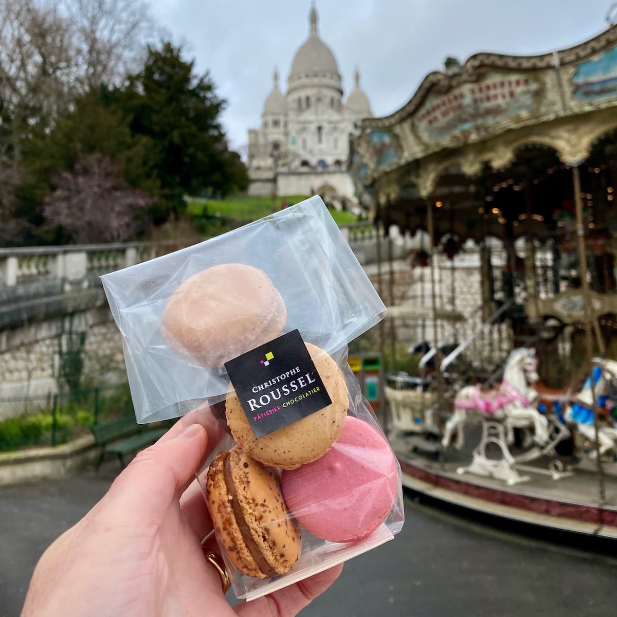 holding a pack of Parisian macarons in front of a carousel and Sacré Coeur basilica in Paris