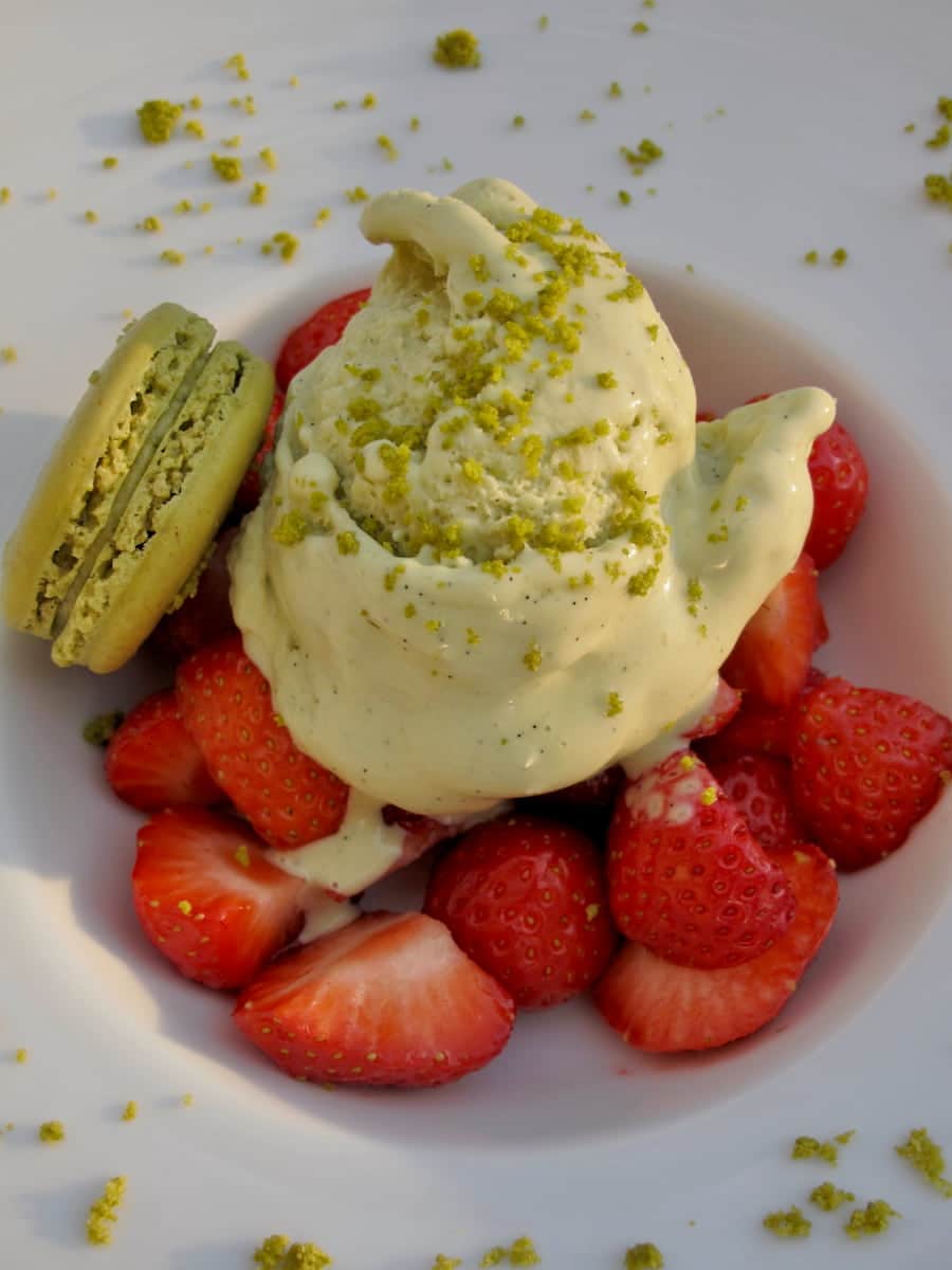 large scoop of pistachio ice cream topped with ground pistachios served with strawberries and a pistachio macaron