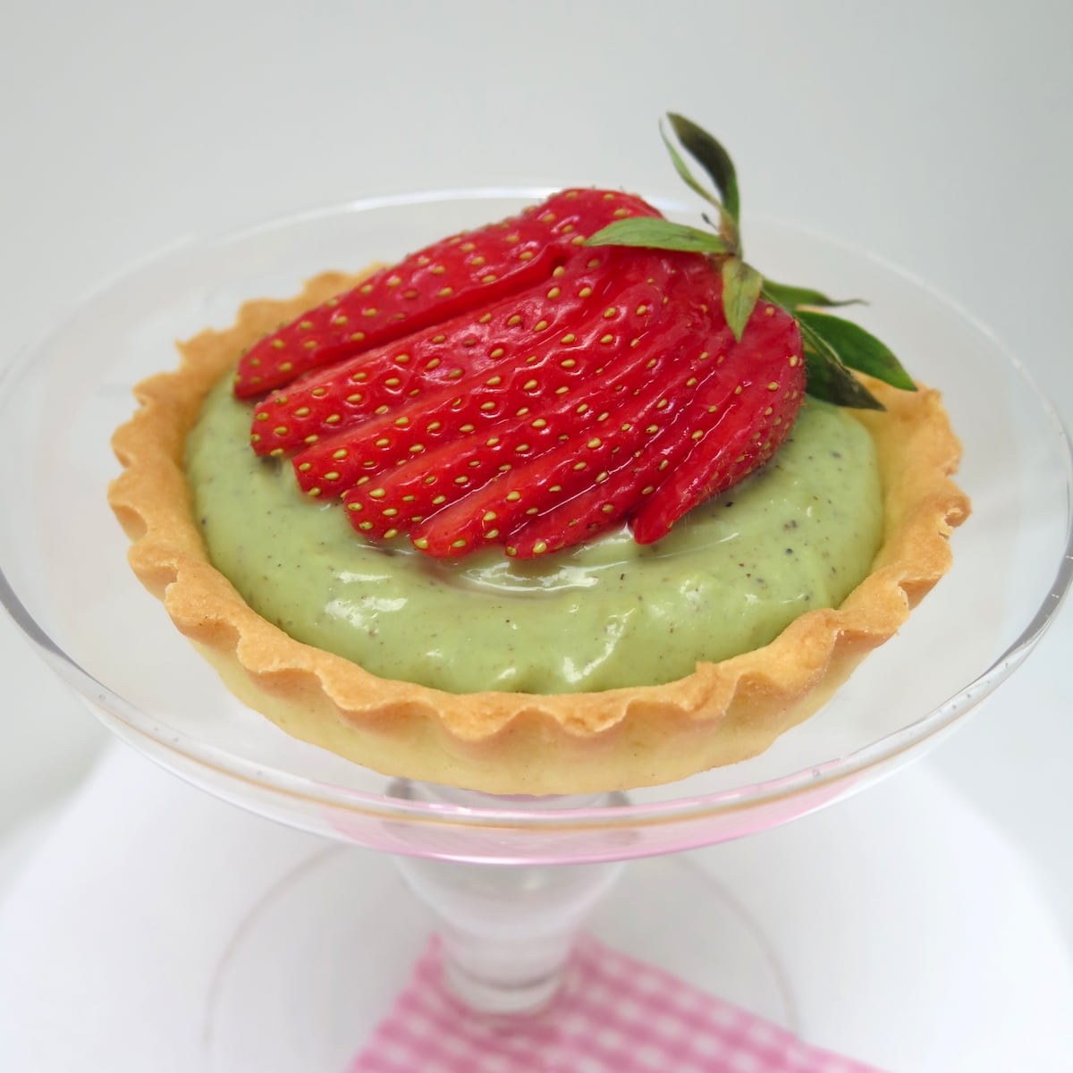 glass dish holding a tart with green pistachio cream topped with a fanned strawberry