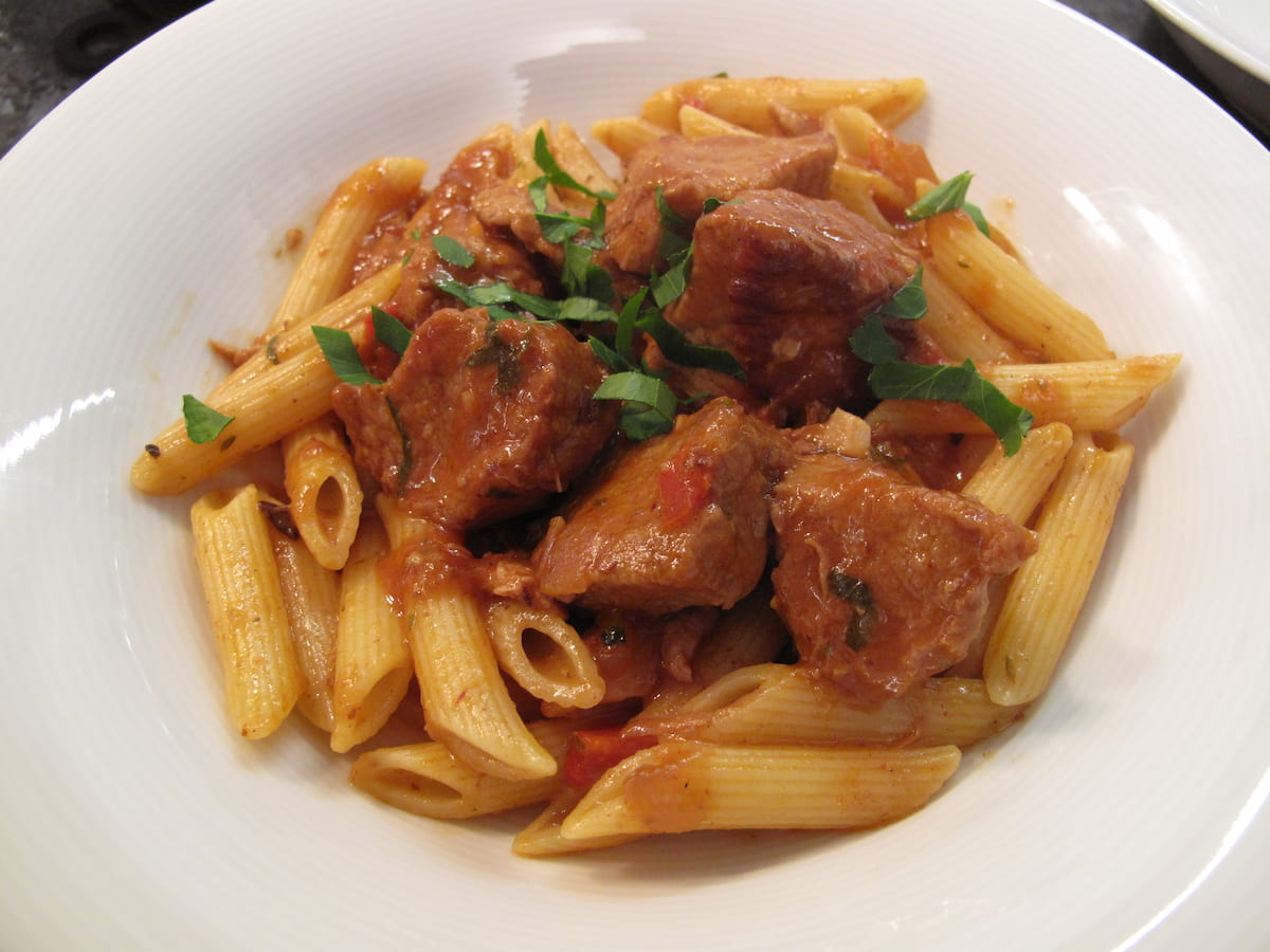 bowl of penne pasta topped with a meat stew speckled with bell peppers