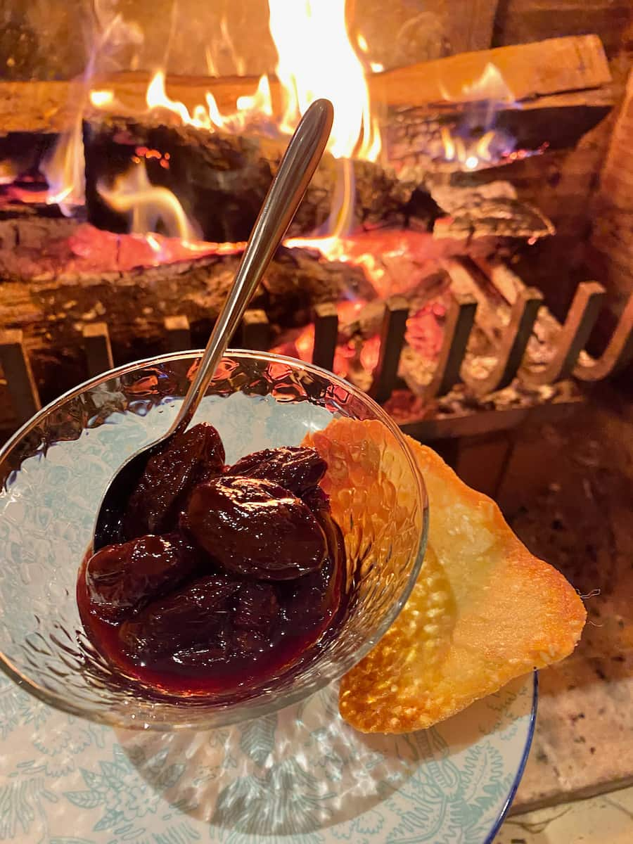 glass bowl of shiny dark prunes in red wine with a crispy tuile cookie in front of a crackling fire