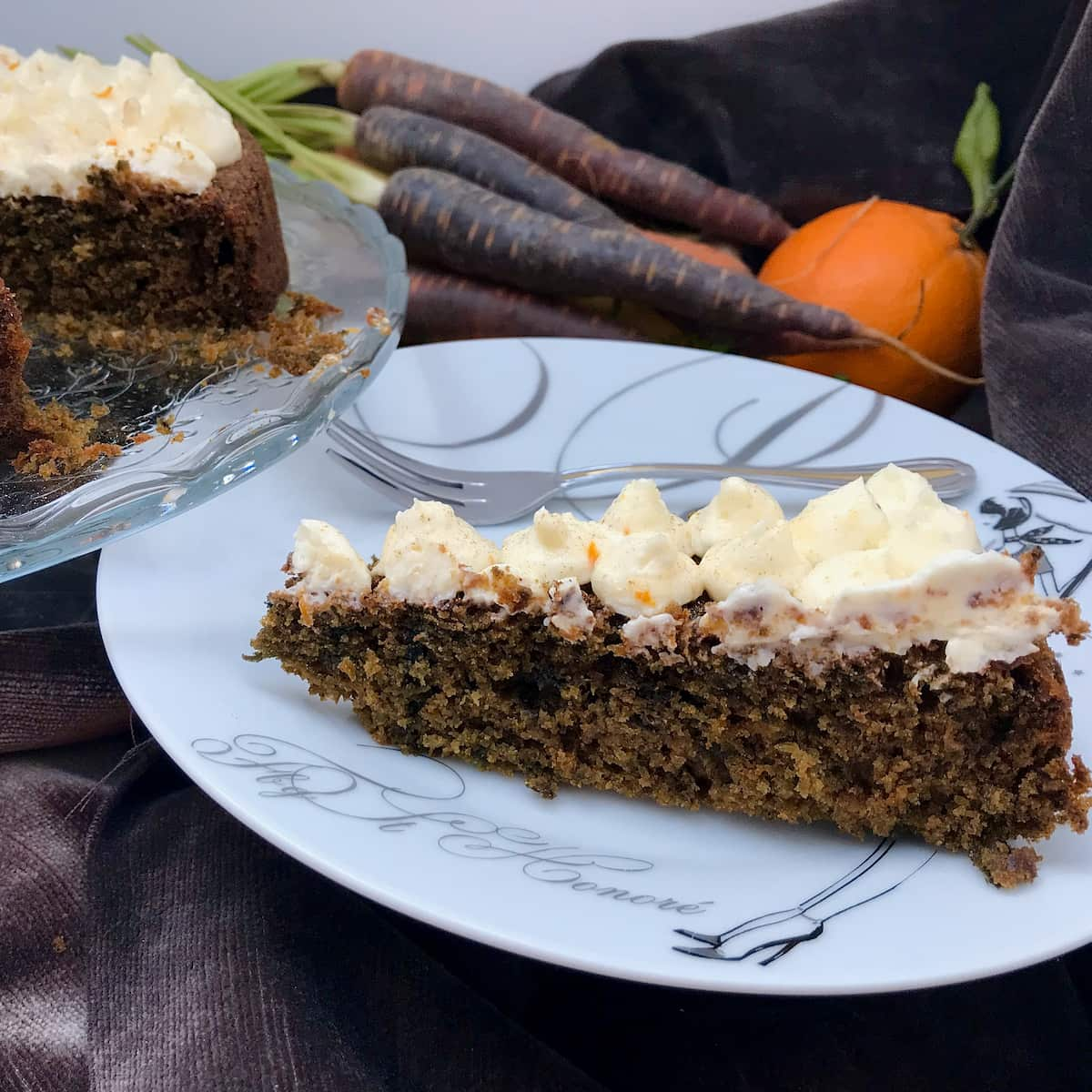 slice of moist, dark cake topped with cream cheese frosting with purple carrots and orange in background