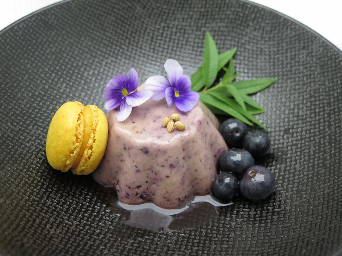 purple moulded dessert on black plate with yellow lemon macaron and shiny blueberries