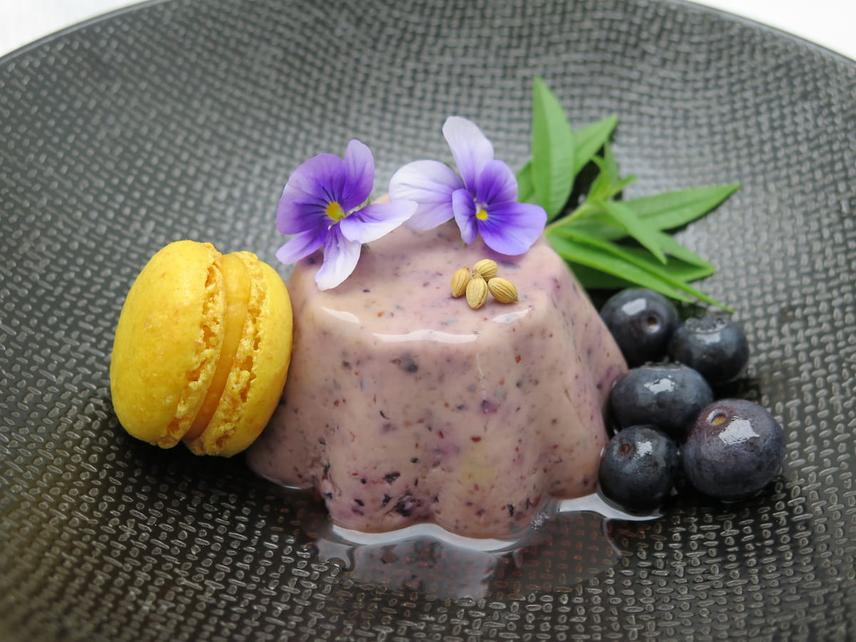 upturned moulded purple dessert with edible flowers, yellow macaron and shiny blueberries