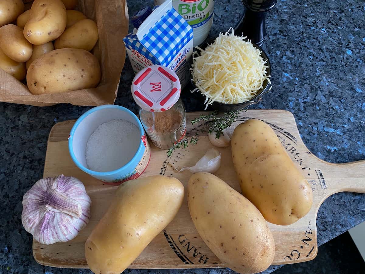 potatoes, garlic, cream, milk, grated cheese, nutmeg, salt and pepper all laid out on a wooden board