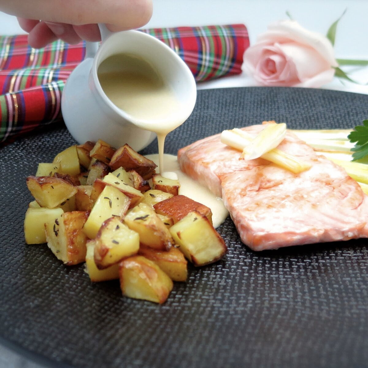 pouring creamy sauce from a mini porcelain jug over crispy potatoes and salmon on black plate