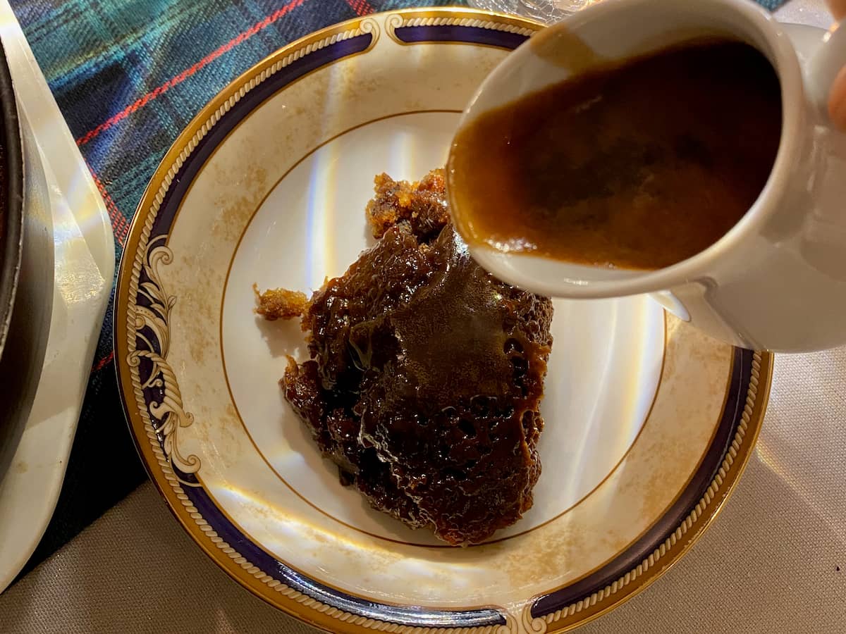 bowl of dark toffee pudding pouring over a little jug of lush sauce