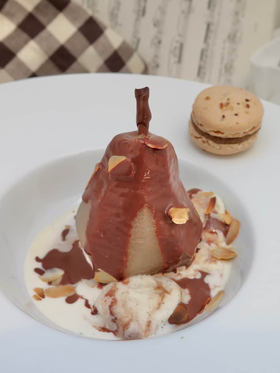 poached pear sitting upright in a bowl topped with chocolate sauce in a pool of ice cream next to a macaron