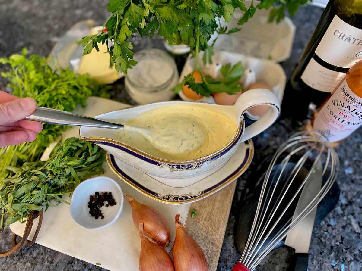 taking a spoon of creamy sauce from a dish, surrounded by herbs, eggs, salt, wine, whisk and pepper