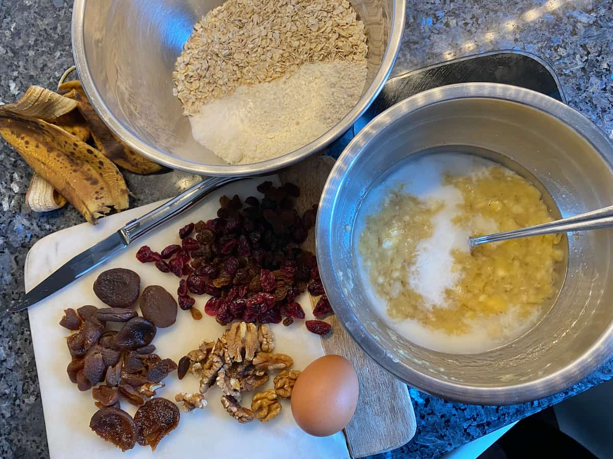 2 bowls containing both dry and wet ingredients, next to dried sultanas, cranberries, apricots and walnuts and banana