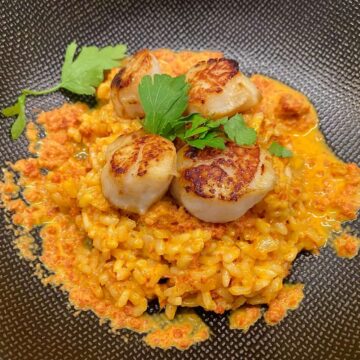 creamy orange coloured rice dish on black plate topped with golden fried scallops with parsley