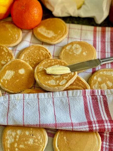 golden little thick pancakes in a tea towel with one being spread with butter