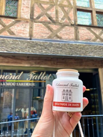 holding a ceramic pot of mustard in front of a historical timbered shop