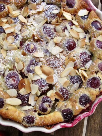 Baked cherry clafoutis dessert in a round dish topped with powdered sugar and flaked almonds