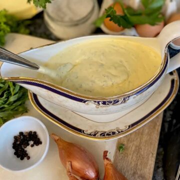 sauce boat of creamy thick sauce with chopped herbs surrounded by shallots, black peppercorns, fresh tarragon and chervil, salt, eggs and a whisk