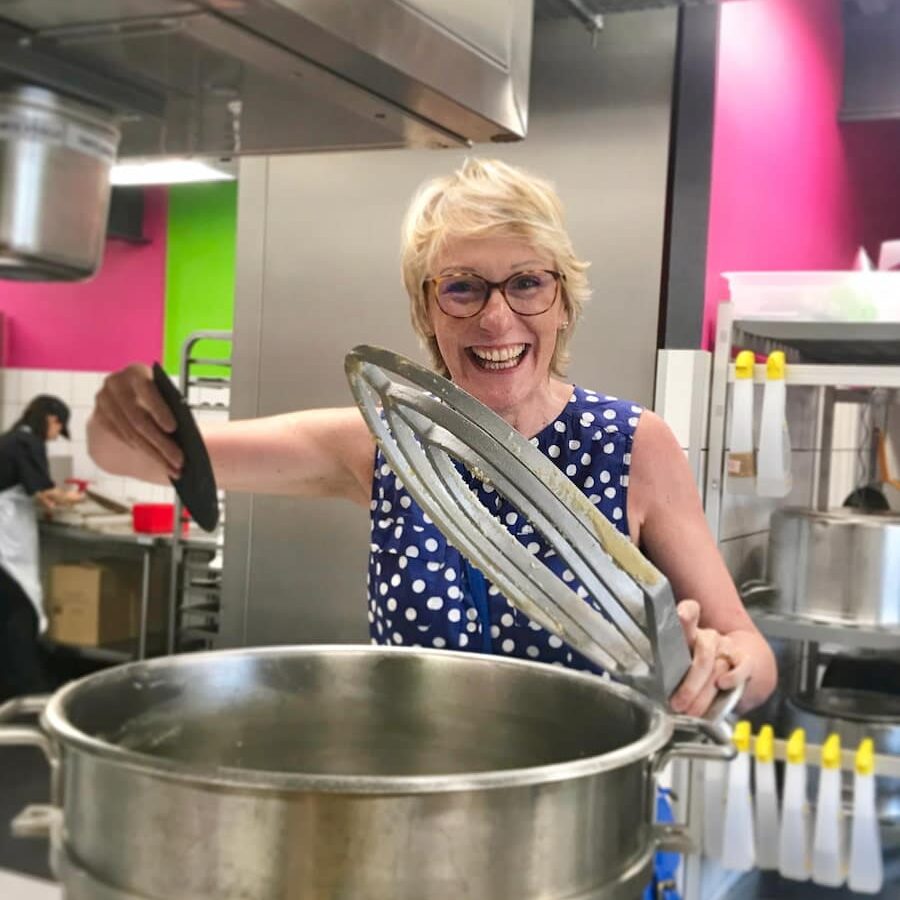 Jill Colonna standing in a French patisserie lab holding a giant whisk over an oversized mixer bowl