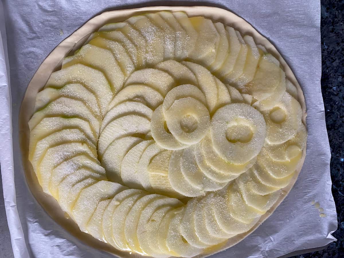 thinly slices apples in a spiral shape on a circle of puff pastry and topped with melted butter and sugar