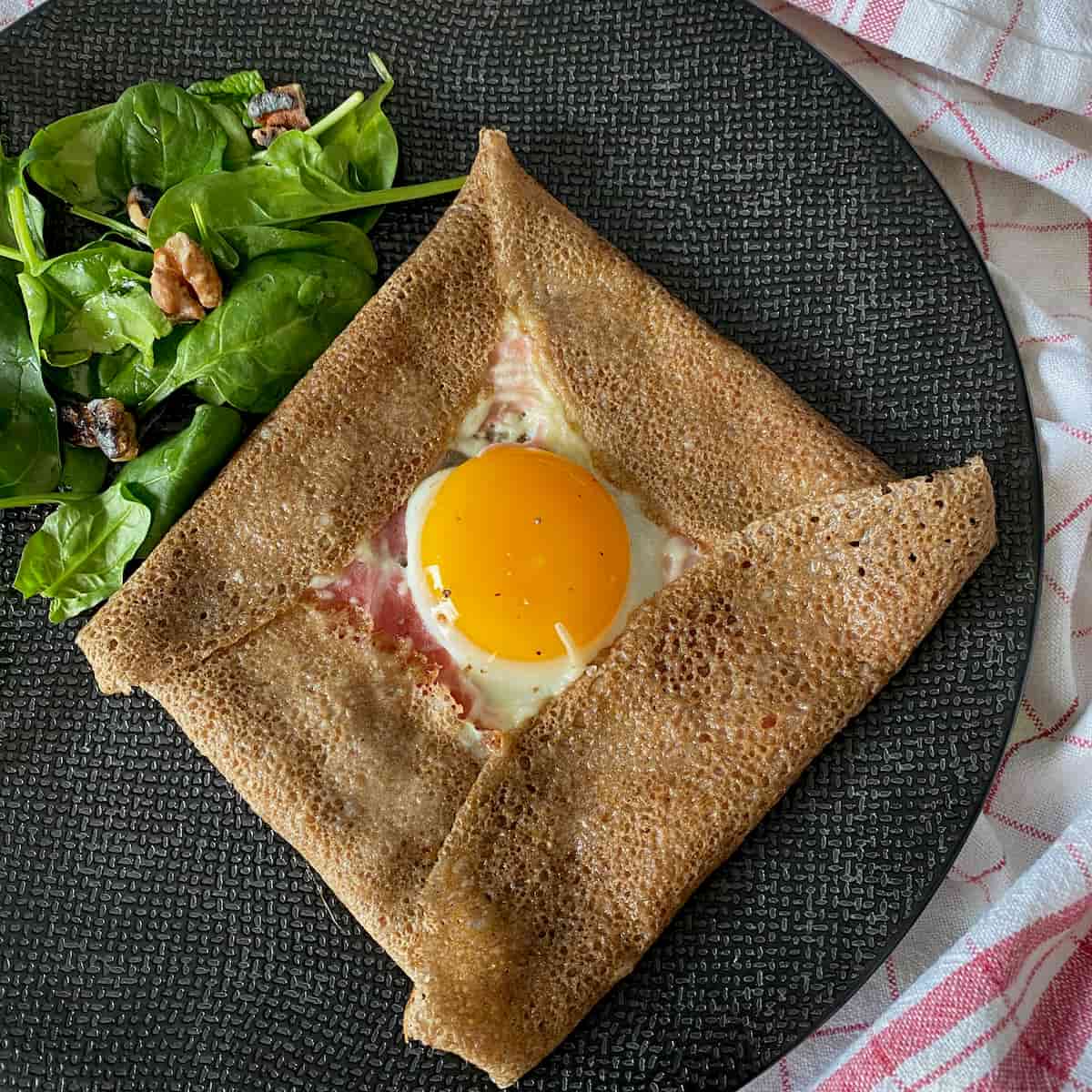thin brown pancake folded into a square with egg in centre, ham and cheese and side salad