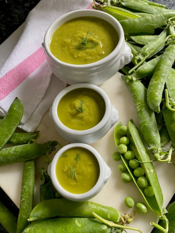 three bowls of bright green soup surrounded by fresh peas and pods