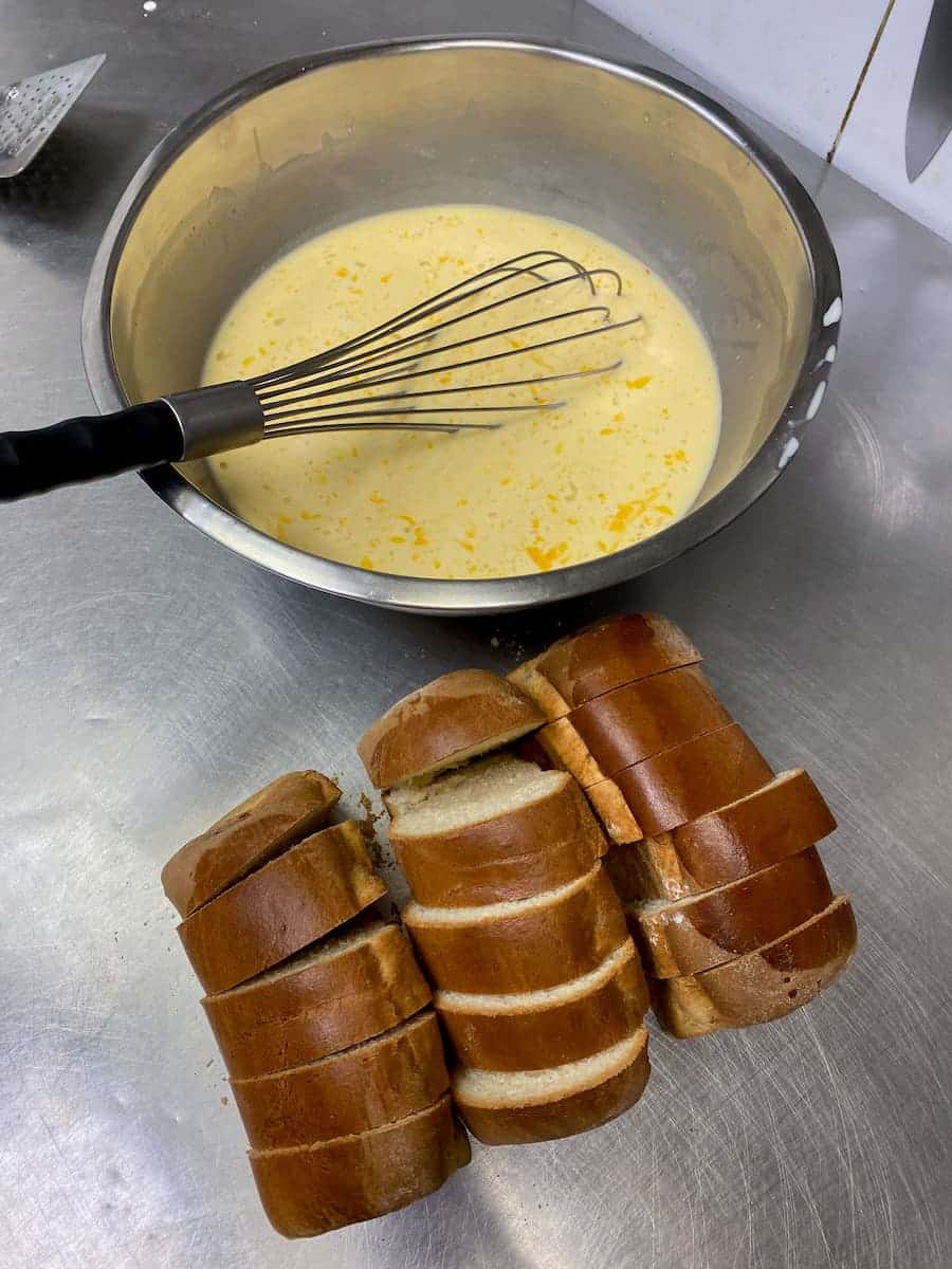 thickly sliced loaves of bread next to a large bowl with eggy mixture and a large whisk