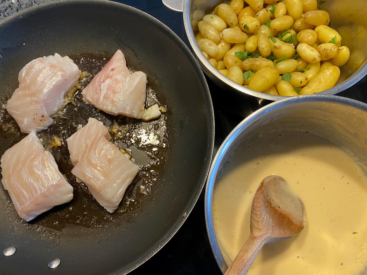 frying fish fillets next to a pan of beurre blanc sauce and another with new potatoes