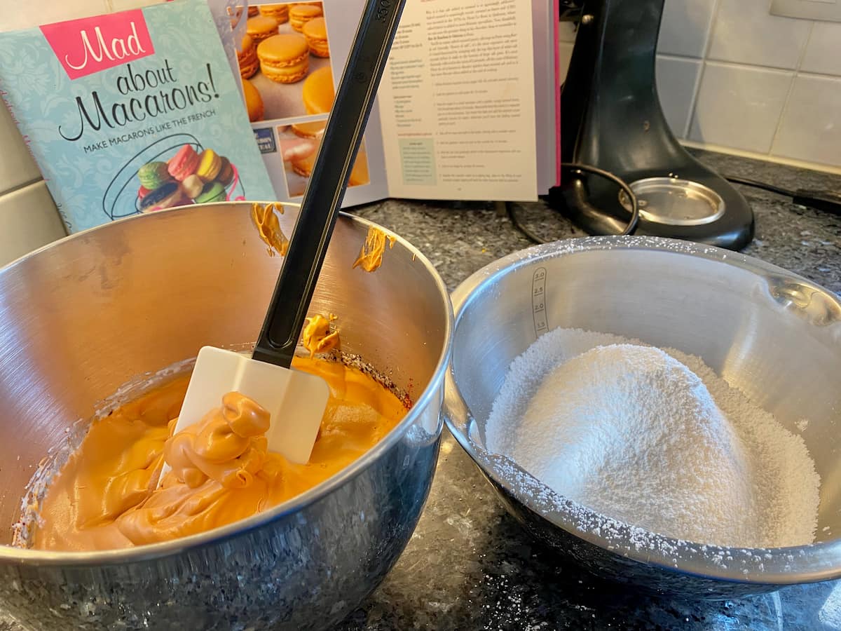 a bowl filled with almond flour and powdered sugar and another with caramel coloured whipped egg whites in front of a macaron recipe book by Jill Colonna