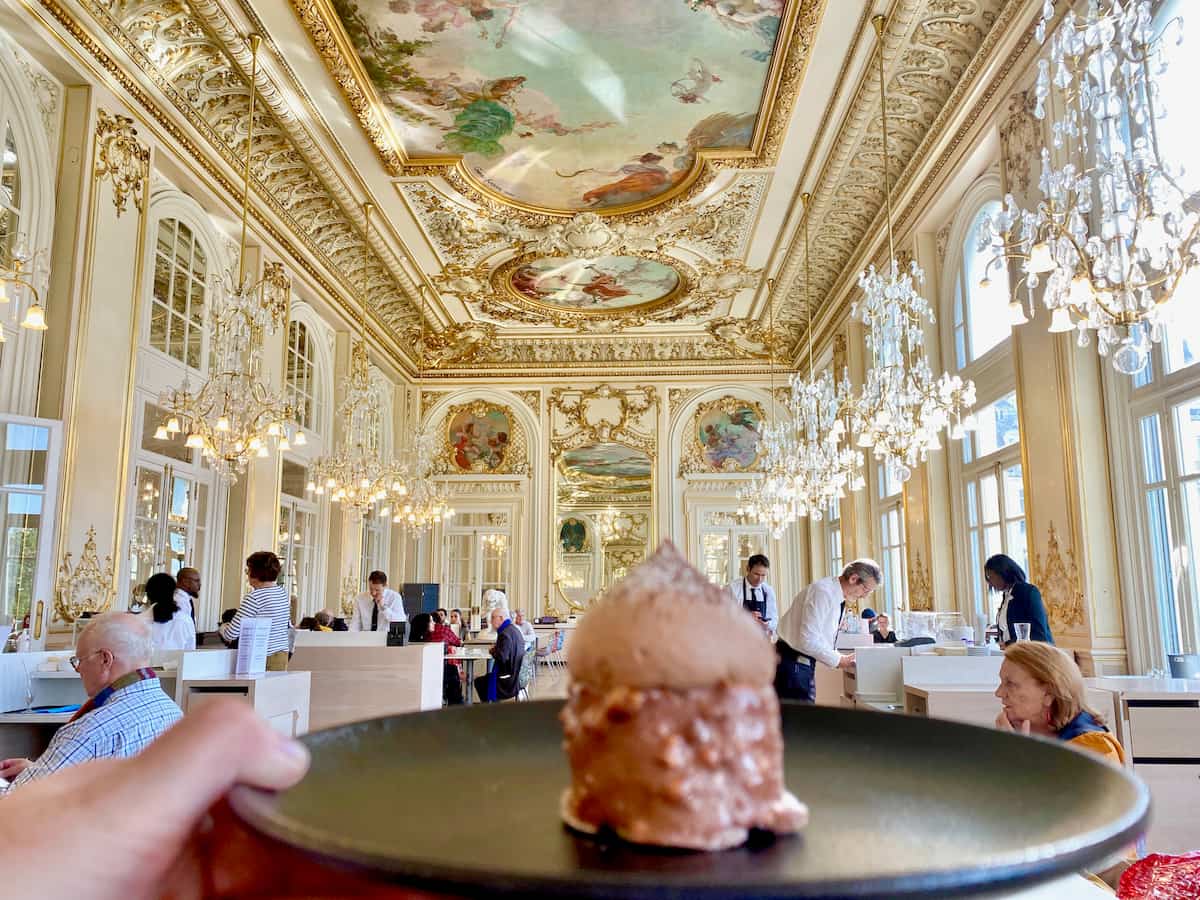 holding a plate with chocolate cake in front of ornate frescoes, gilding and chandeliers in Paris