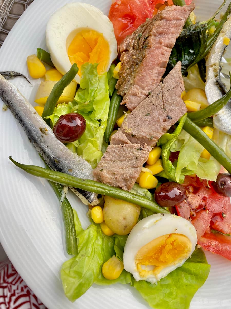 close-up of a composed salad of fresh tuna steak, anchovies, tomatoes, lettuce, olives and boiled eggs but with cooked beans, potatoes and sweetcorn 