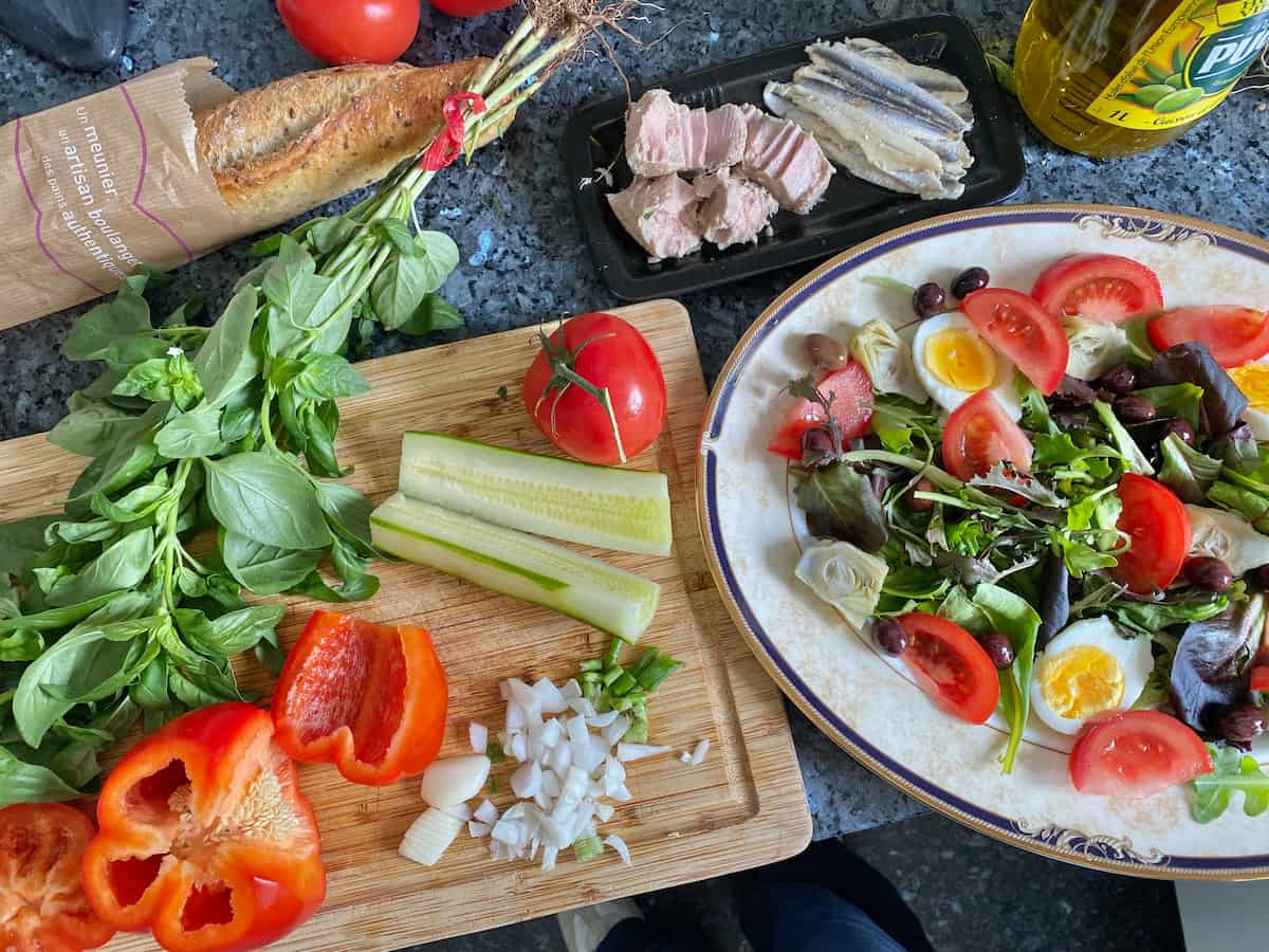 Assembling a large plate of all the ingredients that make up a French salad Nicoise along with a bunch of basil leaves and French baguette
