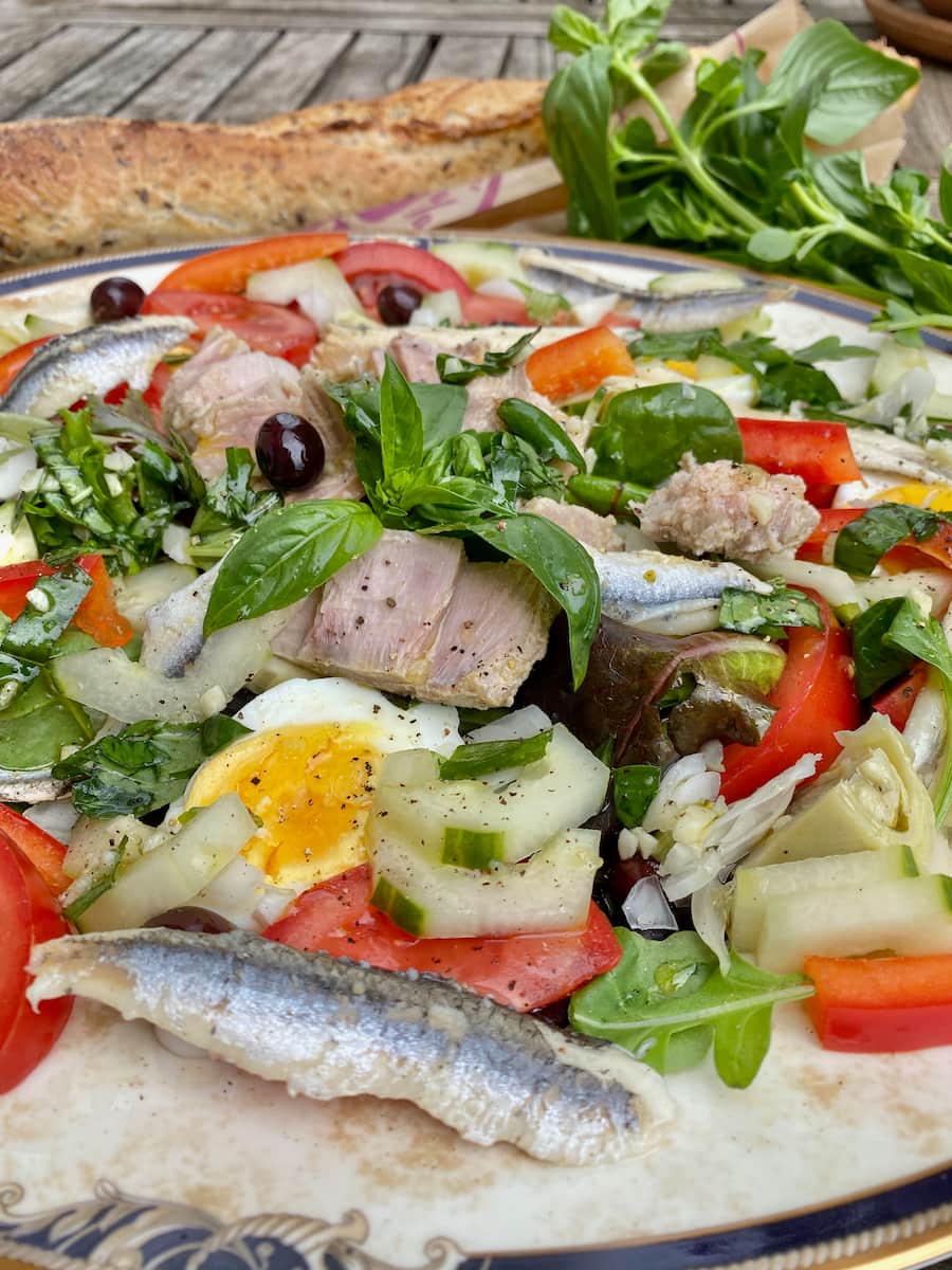 close up of a tuna salad with basil leaves, small black olives, anchovies, tomatoes, cucumber, artichokes, peppers, boiled eggs and garlic.