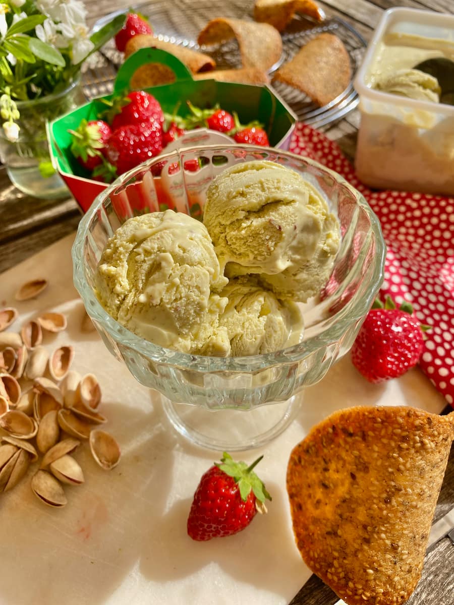 three scoops of pistachio green ice cream in a glass bowl surrounded by fresh strawberries and tuile cookies