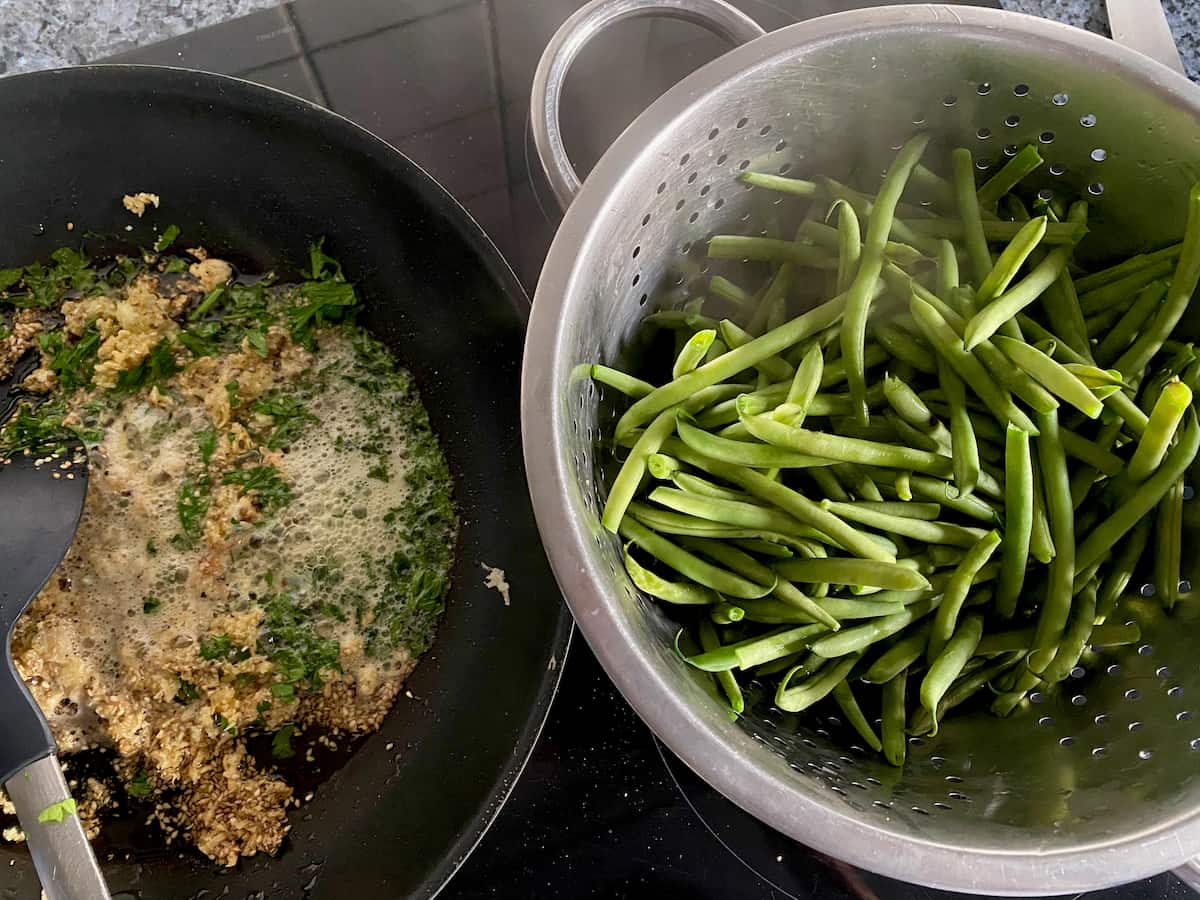 drained green beans in a colander next to a hot pan with the sauté mixture
