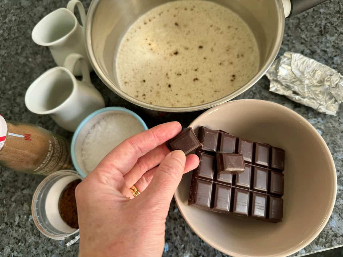 breaking dark bittersweet chocolate tablet into pieces next to a saucepan of milk and vanilla
