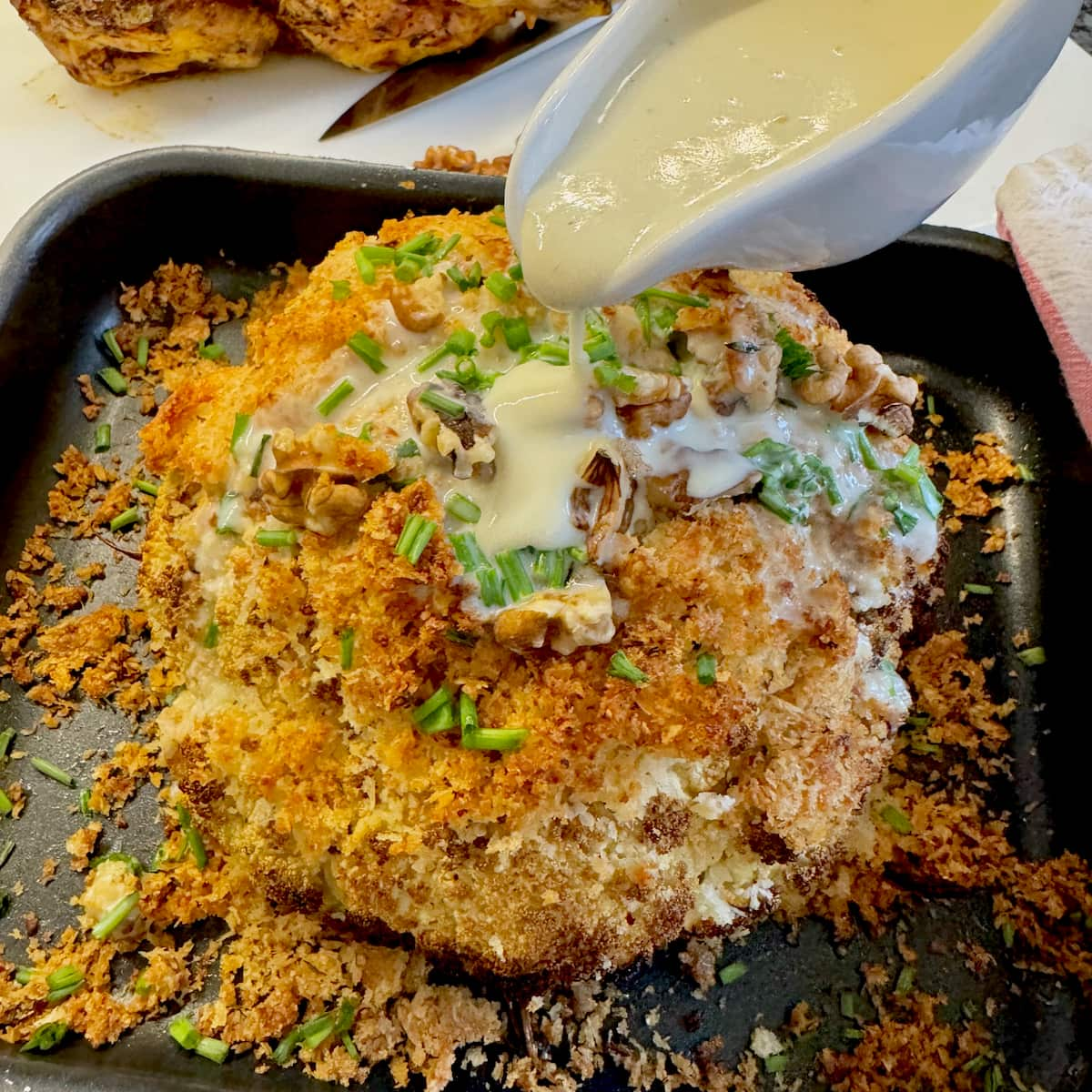 pouring cheese sauce over a whole roasted cauliflower topped with crispy cheese and walnuts