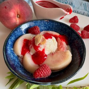 shallow bowl of white poached vanilla peaches topped with homemade raspberry puree and vanilla ice cream with some fresh lemon verbena leaves