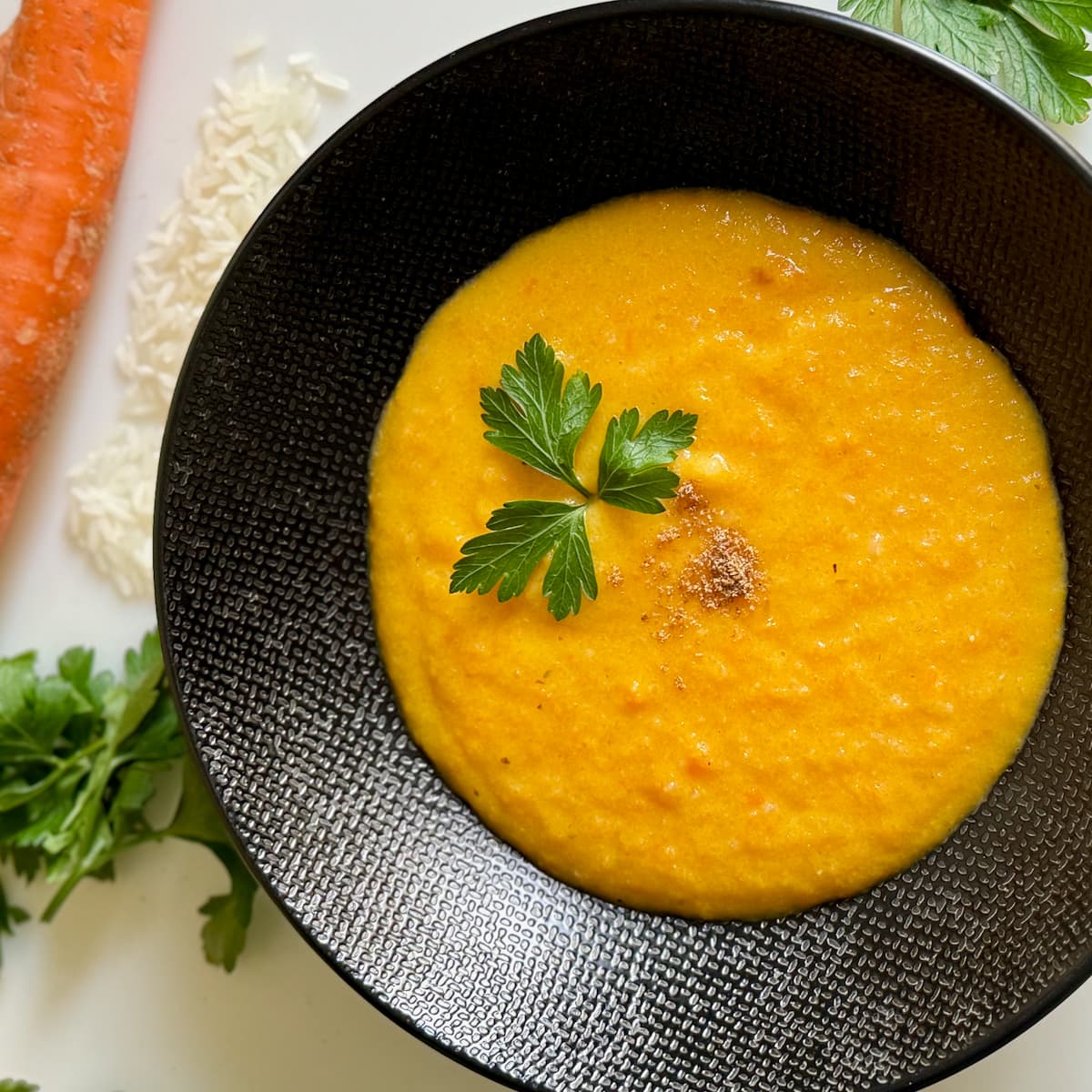 black bowl with a thick and creamy carrot soup, thickened with rice and topped with a sprig of fresh parsley and pinch of ground coriander