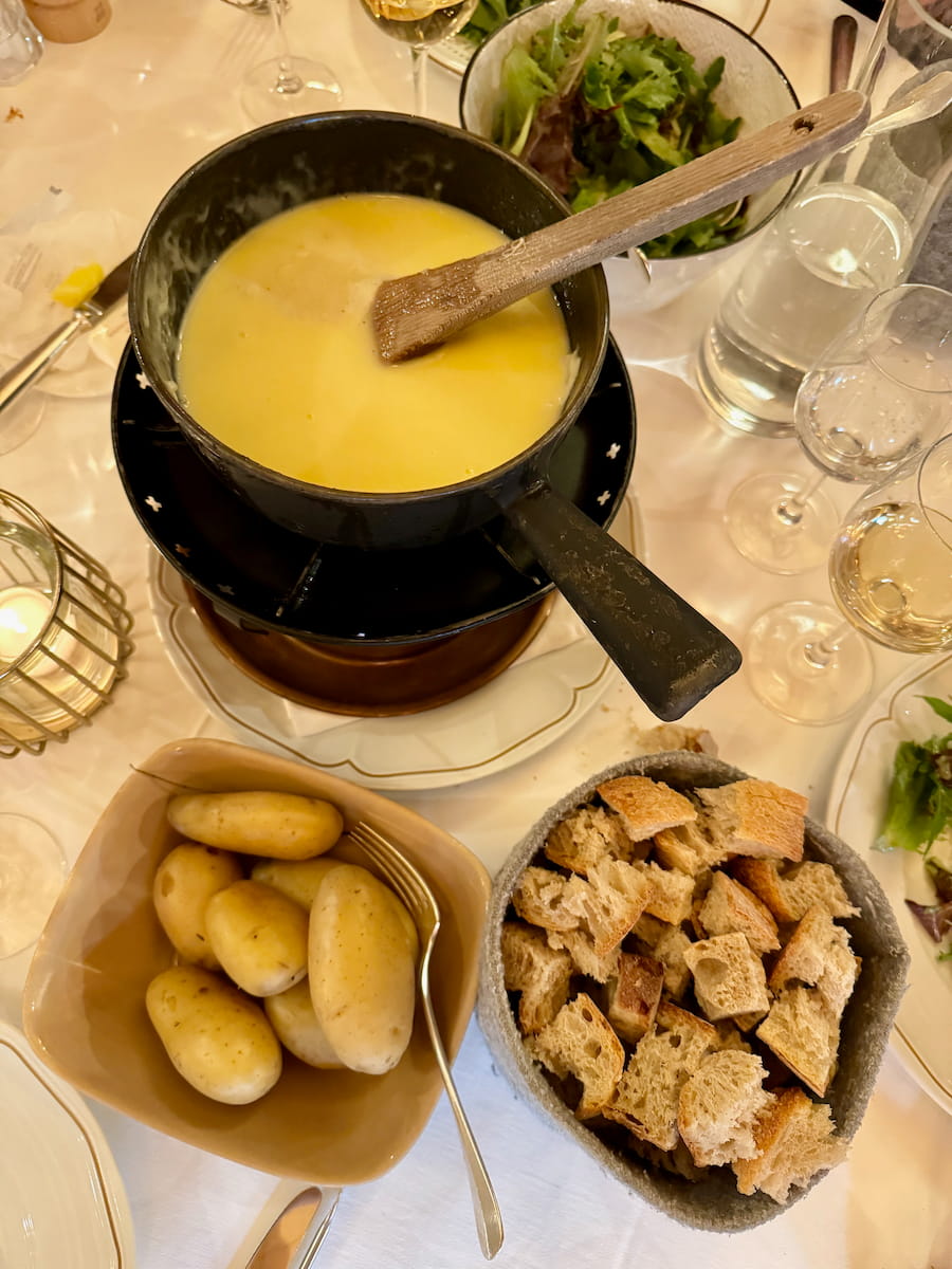 fondue pot heating at the table with a wooden spatula next to a bowl of cubed bread, cooked potatoes in their skins, a green salad and white wine