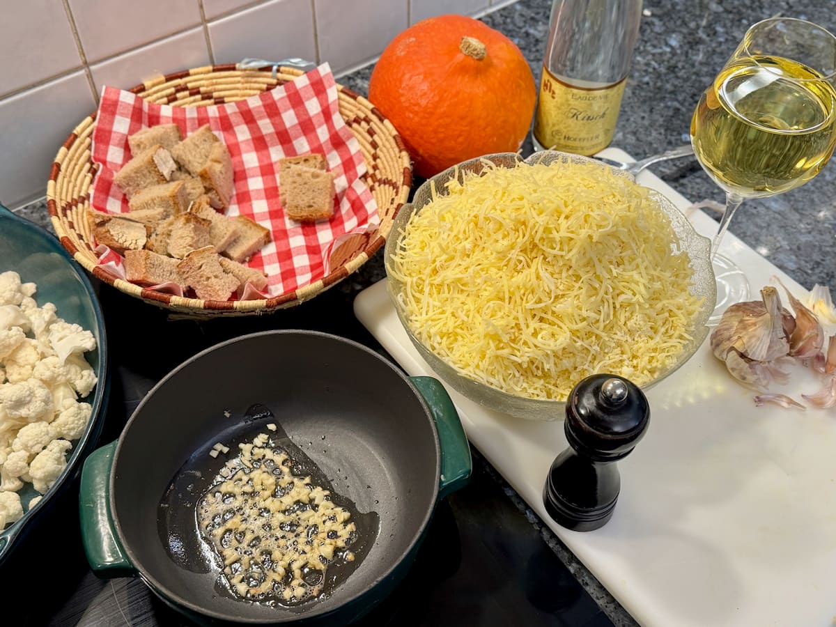 ingredients laid out for a french cheese fondue: grated cheeses in a bowl, chopped garlic frying in butter in a fondue pot next to a large glass of white wine, Kirsch and a bowl of cubed bread