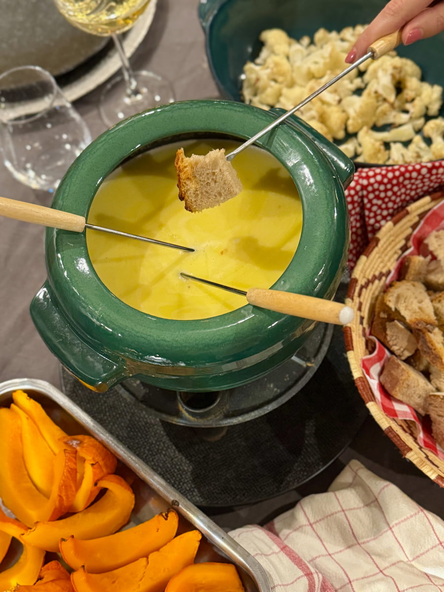 dipping bread on a skewer into a pot of melted cheese next to dishes of roasted cauliflower florets and slices of roasted pumpkin
