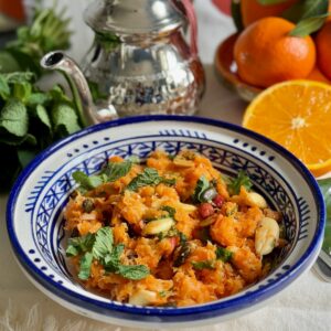 Moroccan handpainted bowl filled with a raw carrot salad topped with fresh mint, toasted nuts and seeds next to oranges and a Moroccan teapot