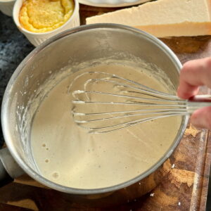whisking a hot parmesan cream sauce in a saucepan next to a block of Parmigiano Reggiano cheese