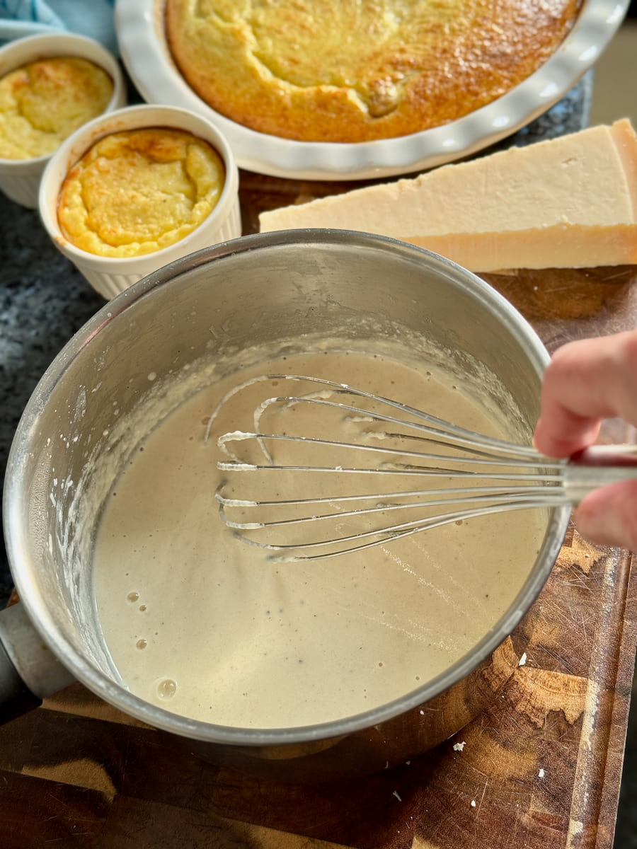 whisked creamy parmesan sauce in a saucepan next to Parmigiano Reggiano dishes of asparagus clafoutis