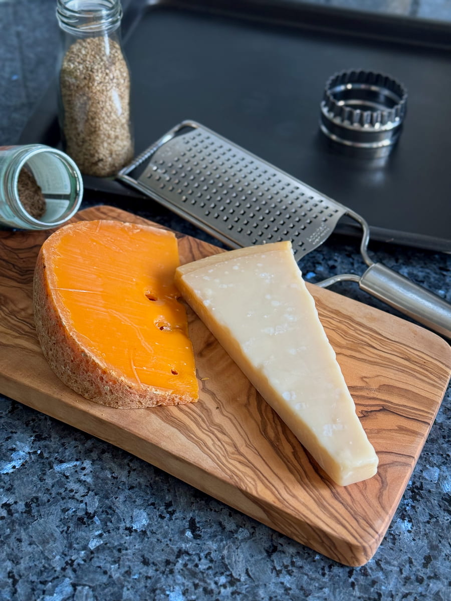 a block of parmesan and aged Gouda cheese on a wooden board next to a grater, baking sheet and cookie cutter with a jar of seeds
